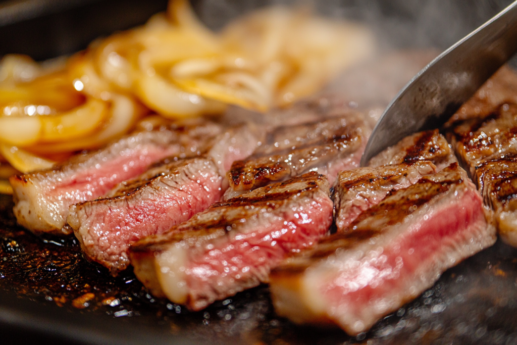 Thinly sliced ribeye steak and caramelized onions cooking on a flat-top griddle.